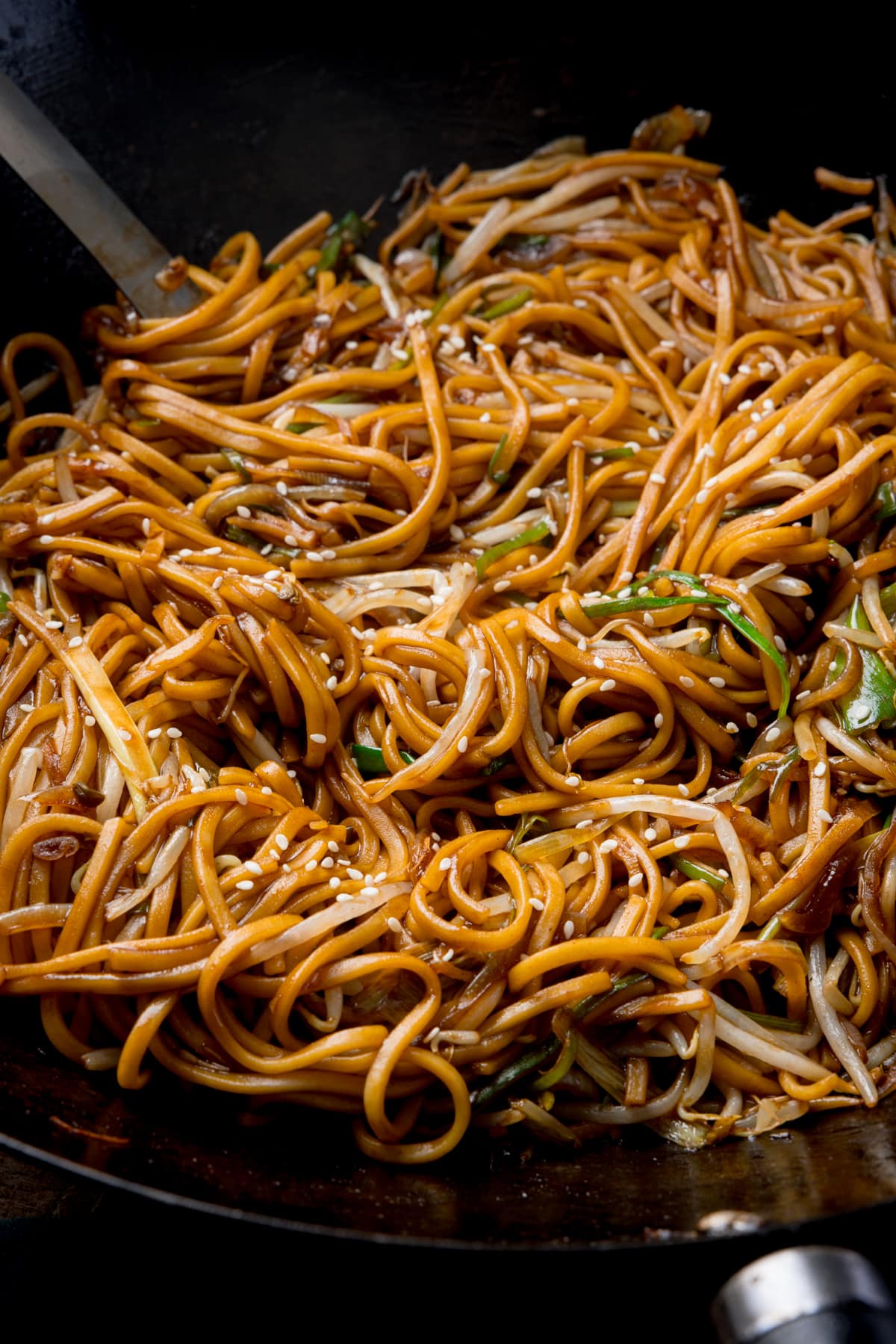 Close-up tall image of stir-fried noodles with beansprouts in a wok, with sesame seeds sprinkled on top.