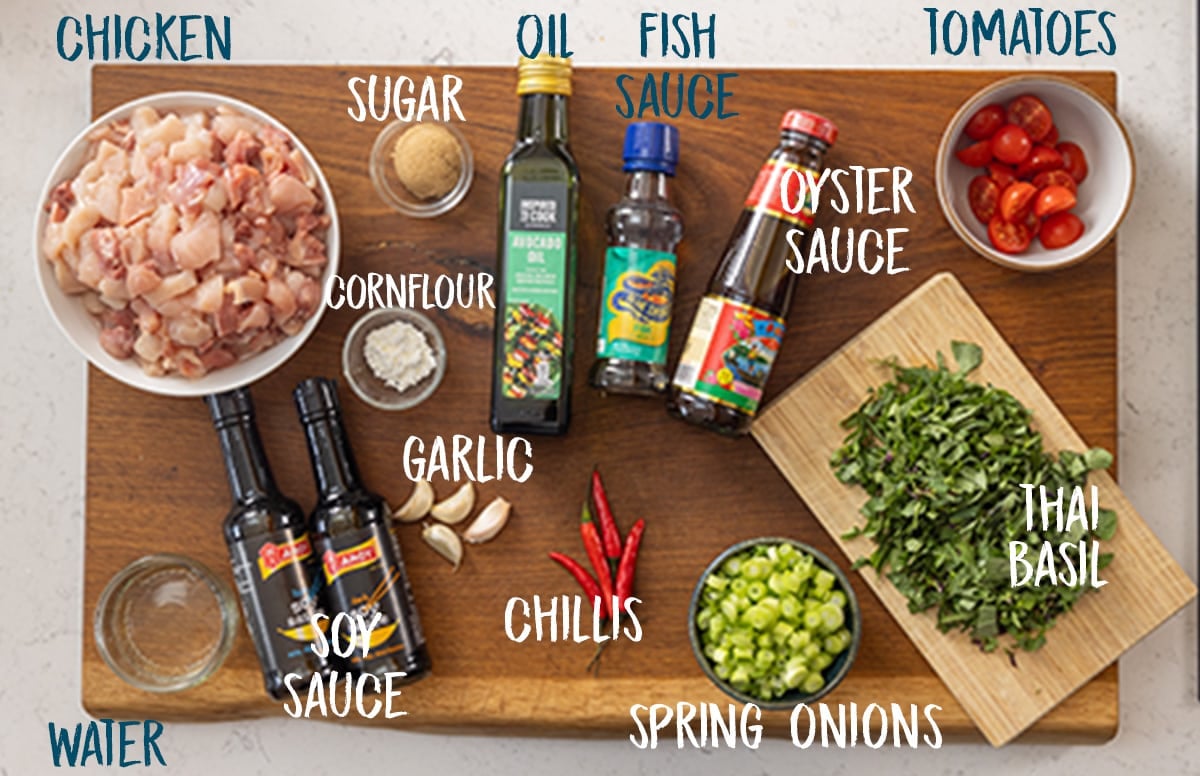 A wide overhead image of the ingredients for thai basil chicken on a wooden cutting board, which is on a white marble surface. The ingredients are labelled in white, except where some ingredient labels flow on top of the white surface, in which case they are blue.