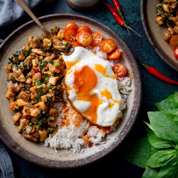 A square overhead image of thai basil chicken served on a grey dish with rice that has a sunny side up egg on top and sliced cherry tomatoes on the top right of the dish. There is a copper fork sticking out of the dish. On the bottom right of the image is a bunch of thai basil. On the top right of the image is the corner of an identical bowl of thai basil chicken. In the background on the top left of the image is a grey handkerchief. there are a few red chillies scattered around. The image is set on a dark blue background.