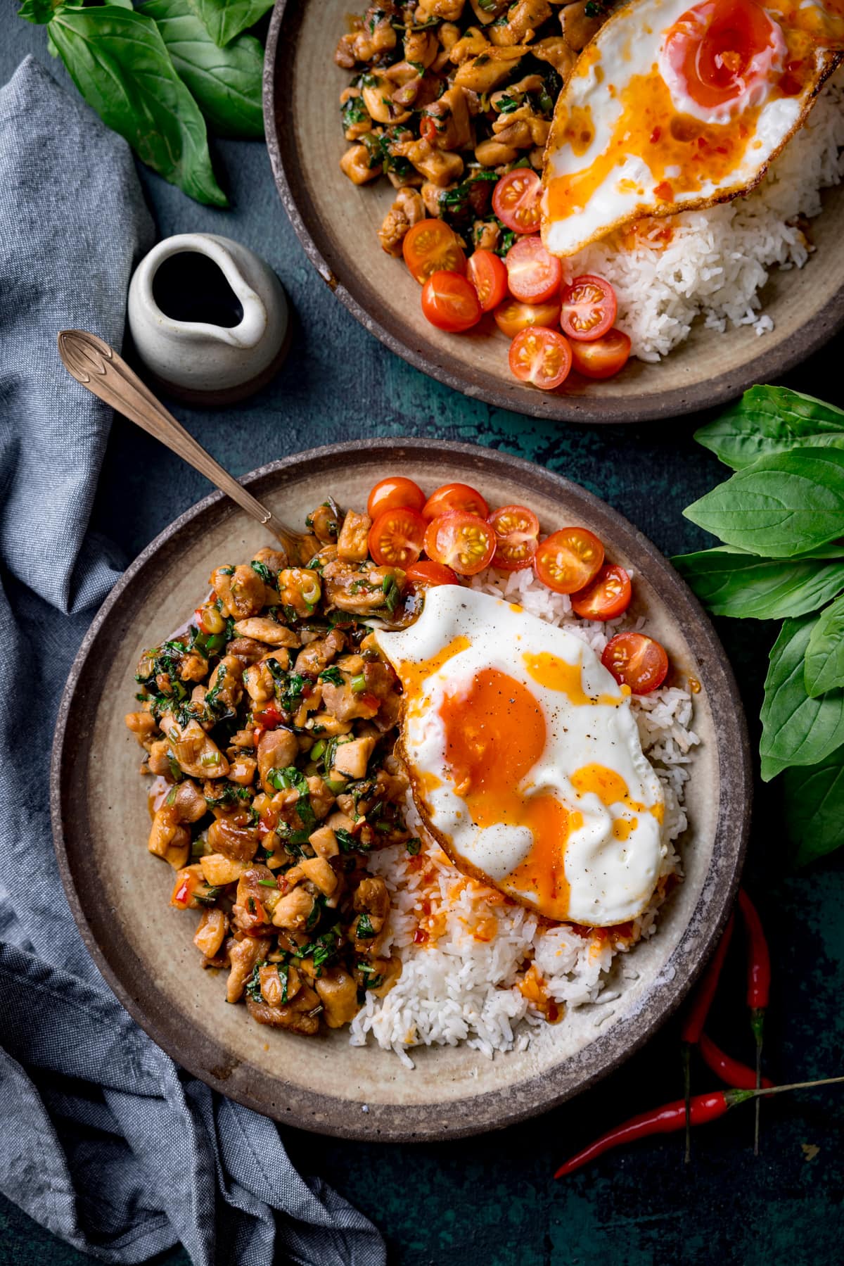 A tall overhead image of thai basil chicken served on a grey dish with rice that has a sunny side up egg on top and sliced cherry tomatoes on the top right of the dish. There is a copper fork sticking out of the dish. On the bottom right of the image is a bunch of thai basil. On the top right of the image is the corner of an identical bowl of thai basil chicken and a small white jug of sauce. In the background on the top left of the image is another bunch of basil and a grey handkerchief. there are a few red chillies scattered around. The image is set on a dark blue background.