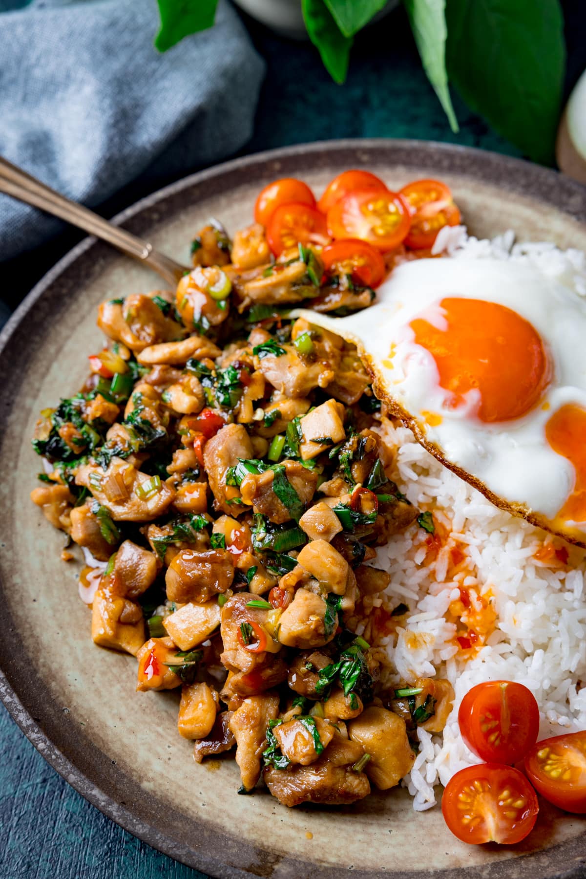 A tall close-up image of thai basil chicken served on a grey dish with rice that has a sunny side up egg on top and sliced cherry tomatoes on the top right of the dish. There is a copper fork sticking out of the dish. In the background on the top left of the image is a grey handkerchief. The image is set on a dark blue background.