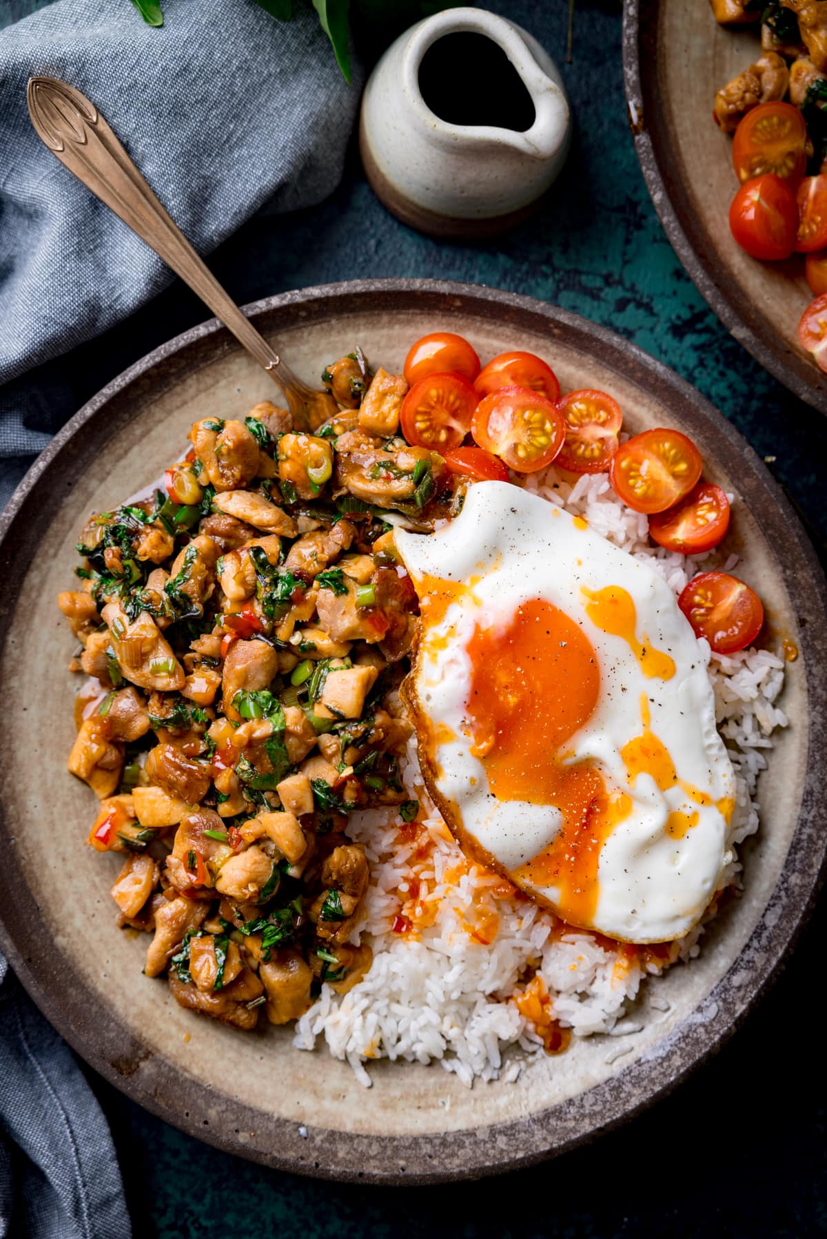 A tall overhead image of thai basil chicken served on a grey dish with rice that has a sunny side up egg on top and sliced cherry tomatoes on the top right of the dish. There is a copper fork sticking out of the dish. On the bottom right of the image is a bunch of thai basil. On the top right of the image is the corner of an identical bowl of thai basil chicken and a small white jug of sauce. In the background on the left of the image is a grey handkerchief. The image is set on a dark blue background.