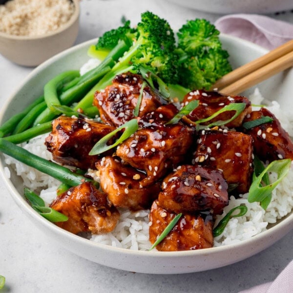 A square photo of Air Fryer Teriyaki Salmon served on white rice, with broccoli, and topped with sesame seeds and spring onions in a white bowl. There is a pair of wooden chopsticks sticking out of the dish to the right. In the top left of the background, there is a cream dish filled with white sesame seeds. On the top right of the background, you can see a white napkin and you can slightly see another white bowl. This is all set on a white surface.