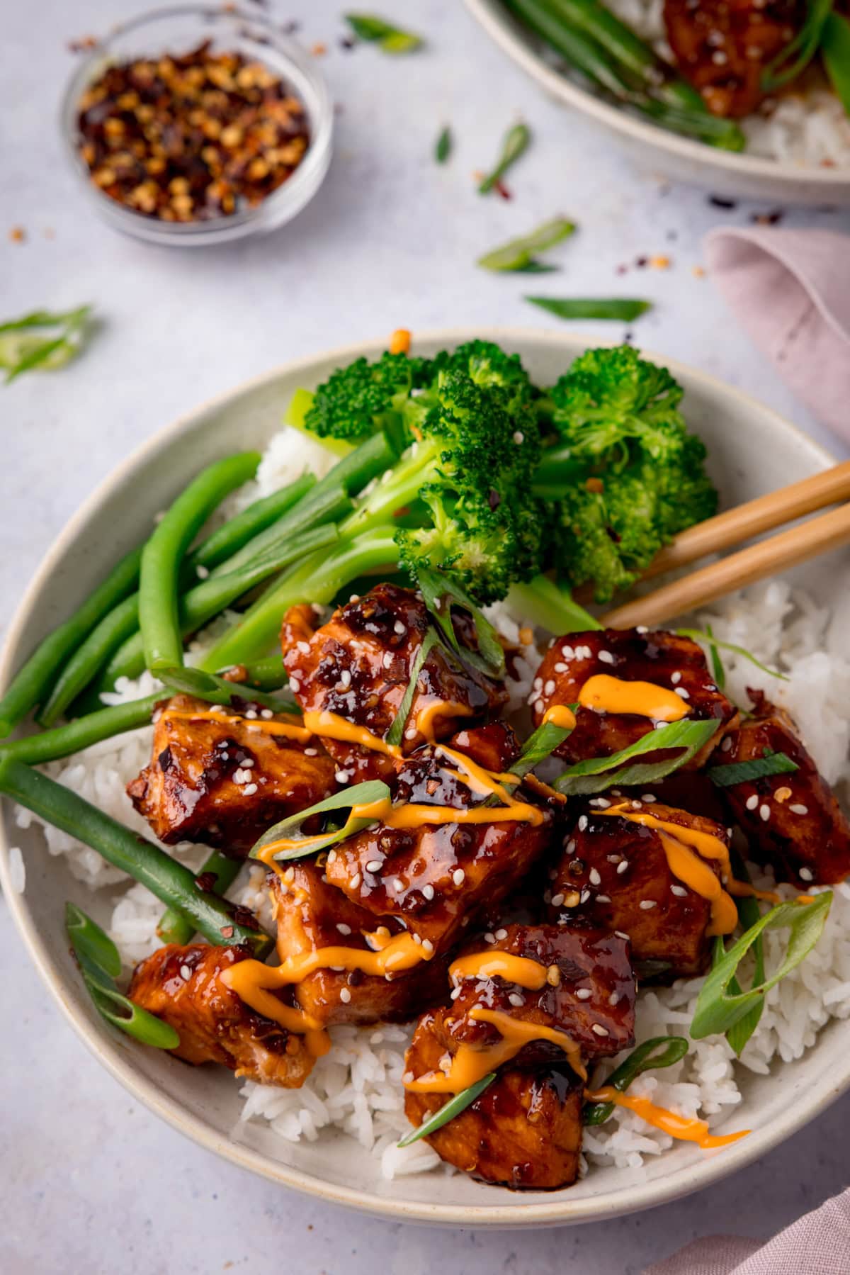 A tall overhead photo of Air Fryer Teriyaki Salmon served on white rice, with broccoli, and topped with sesame seeds and spring onions in a white bowl. There is a pair of wooden chopsticks sticking out of the dish to the right. In the top left of the background, there is a white dish filled with dried chilli flakes. On the top right of the background, you can see a white napkin and you can slightly see another white bowl of Air Fryer Teriyaki Salmon. There are sesame seeds and spring onions scattered around. This is all set on a white surface.