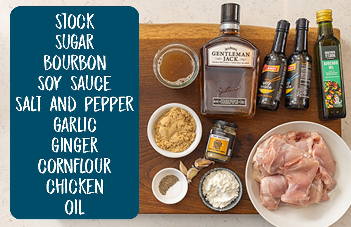 Overhead shot of the ingredients for bourbon chicken on a wooden board, placed on a white counter. On the left of the image is a blue box that has white text, listing the ingredients as follows: stock, sugar, bourbon, soy sauce, salt, pepper, garlic, ginger, cornflour, chicken, and oil.