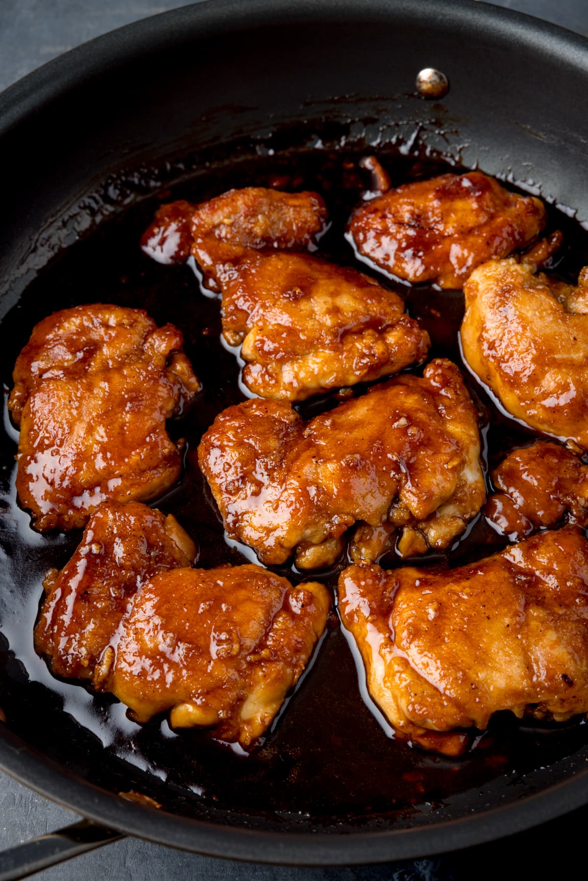 A tall shot of the whole bourbon chicken fillets in a dark grey pan. The pan is resting on a grey background with a dark blue napkin.