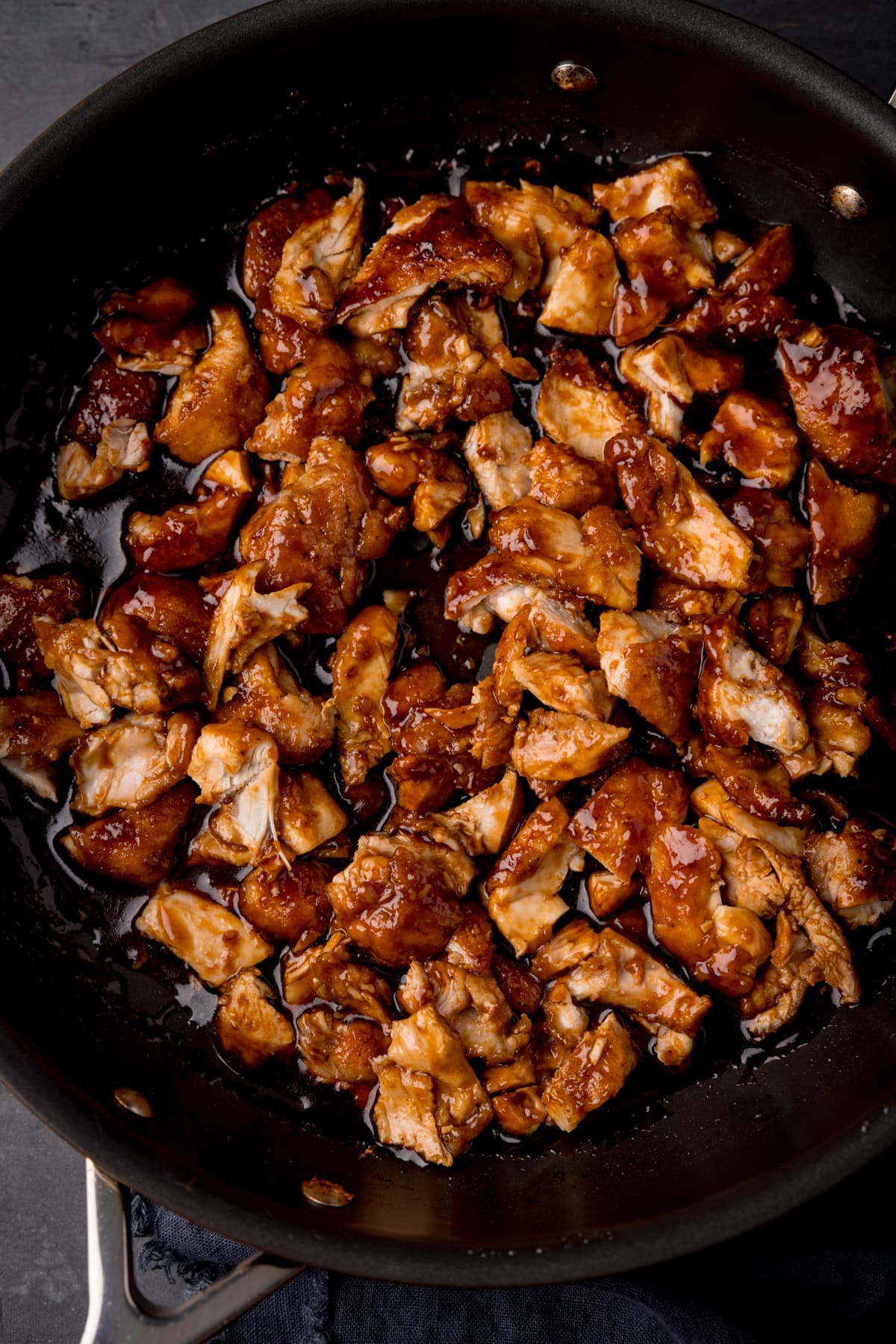 A tall shot of the bourbon chicken in a dark grey pan. The pan is resting on a grey background with a dark blue napkin.