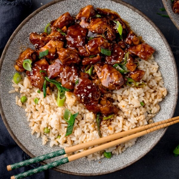 A square, overhead shot of a grey plate of bourbon chicken with egg fried rice, topped with spring onions and sesame seeds. The plate has a pair of wooden chopsticks lying on it. On the left, below the plate of bourbon chicken, is a dark blue napkin. This is all set on a dark blue background.