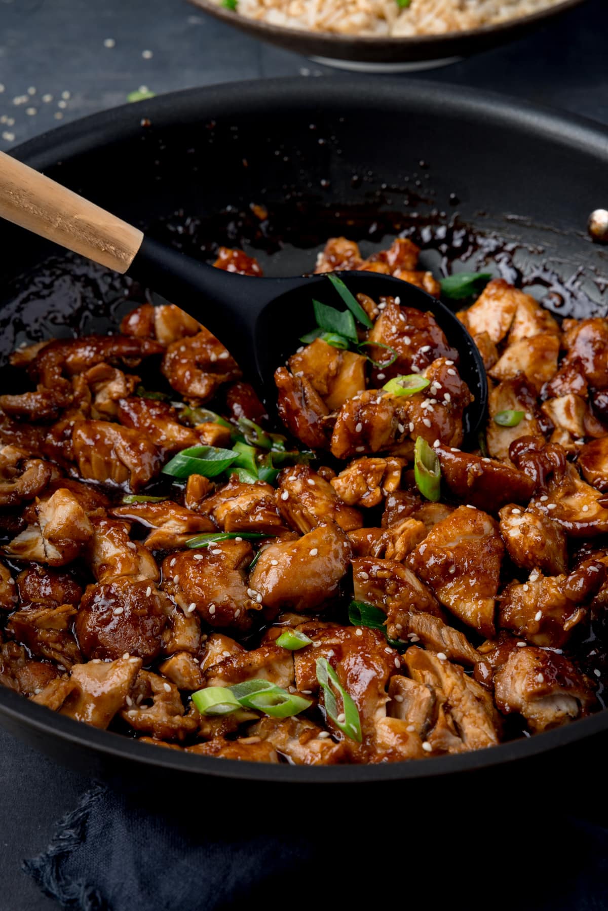 A tall shot of bourbon chicken in a dark grey pan, topped with spring onions and sesame seeds. There is a black spoon with a wooden handle scooping some of the chicken out of the pan. The pan is resting on a grey background with a dark blue napkin. In the top of the image you can slightly see a blue bowl of egg-fried rice.