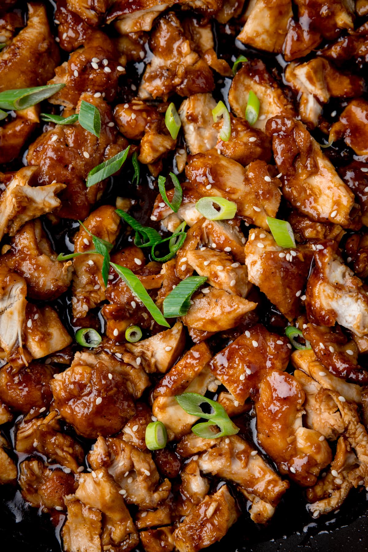 A tall close-up shot of bourbon chicken, topped with spring onions and sesame seeds.