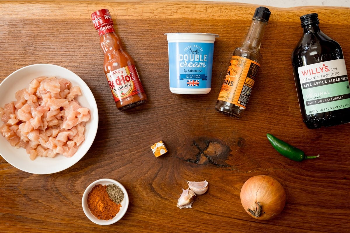 An overhead shot of the filling ingredients are arranged on a wooden cutting board, which is on a white surface. The ingredients are as follows: chicken, hot sauce, double cream, Worcestershire sauce, apple cider vinegar, paprika, pepper, chicken stock cube, garlic, onion and a jalapeno.