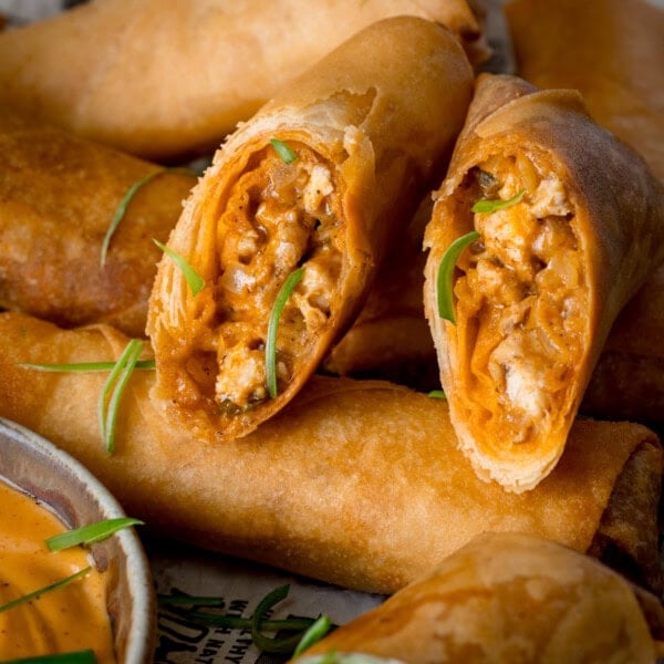 A square image of buffalo chicken spring rolls, topped with small shreds of spring onions. The spring rolls are placed on newspaper and in the bottom left of the image there is a white dish of jalapeno mayonnaise.