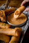 A tall image of a stack of buffalo chicken spring rolls, topped with small shreds of spring onions. In the centre of the image, there is a white dish of jalapeno mayonnaise, there is half of a spring roll dipped into the sauce, and the other half lays beside the dish. The spring rolls are placed on newspaper, on a tray. In the top left of the tray, you can see the wooden handle of a knife. The whole tray is placed on a wooden surface.