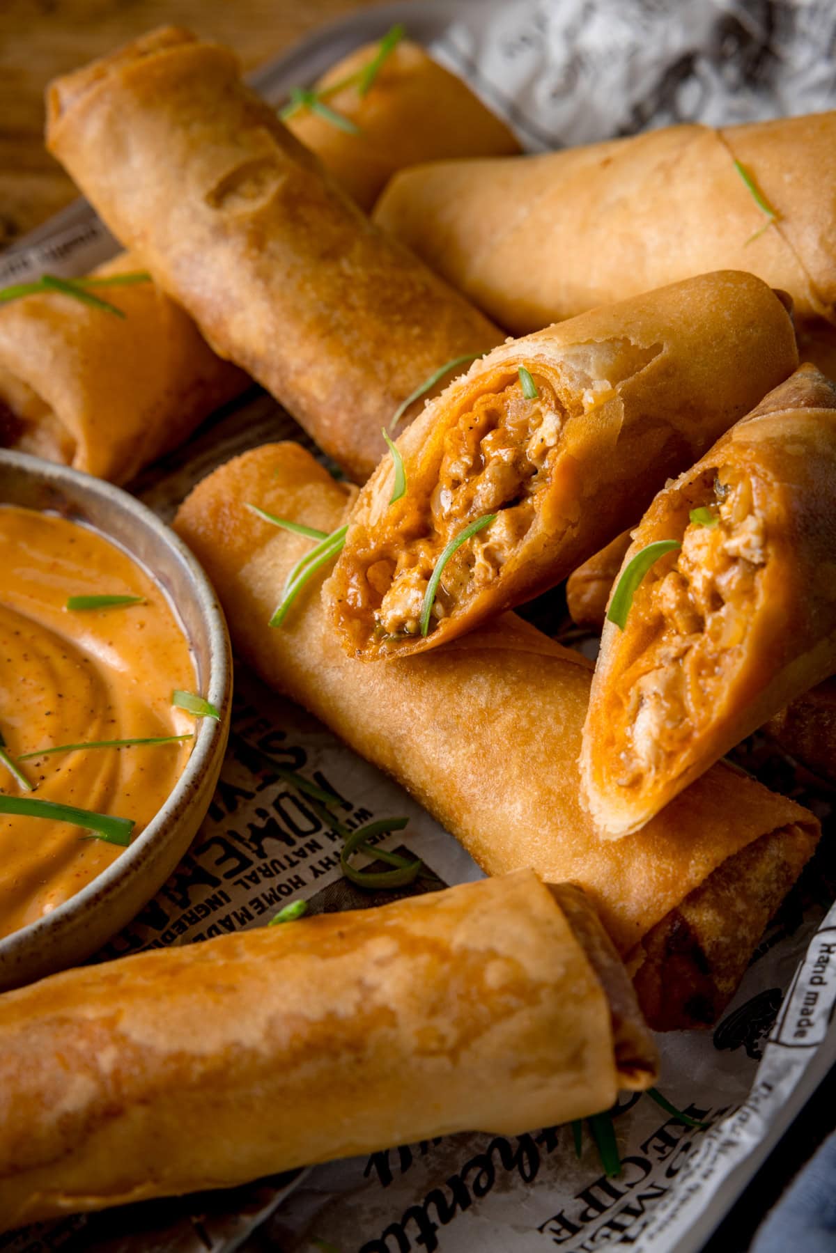 A tall image of a stack of buffalo chicken spring rolls, topped with small shreds of spring onions. In the centre of the image there is two halves of a spring roll, so you can see the inside. The spring rolls are placed on newspaper and in the bottom left of the image there is a white dish of jalapeno mayonnaise.