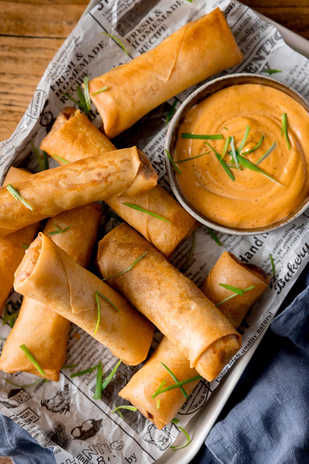 A tall overhead shot of a stack of spring rolls, topped with shreds of spring onions. They are placed on a tray that is lined with newspaper. In the top right of the tray there is a white dish of jalapeno mayonnaise, which also is topped with shreds of spring onions. There is a grey napkin tucked underneath the bottom right of the tray, which is placed on a wooden surface.