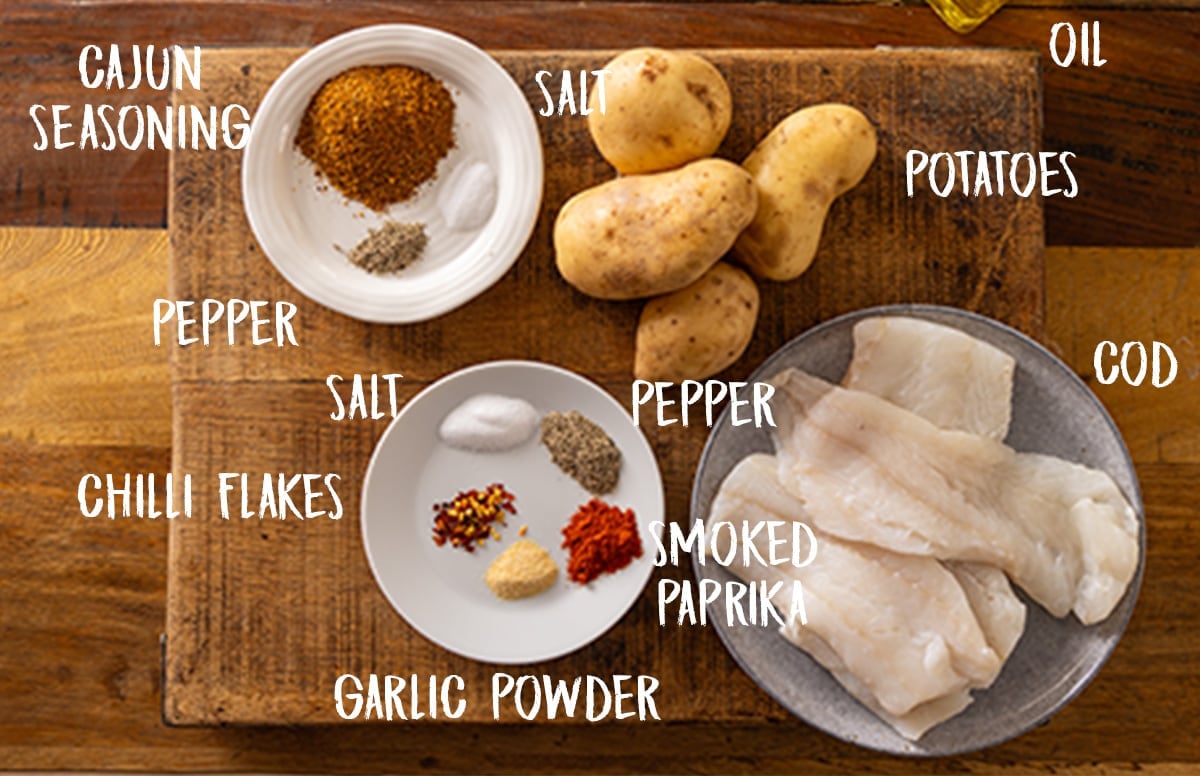 The following ingredients are arranged on a wooden chopping board labelled in white: Potatoes, Cod, Oil, Salt, Pepper, Smoked Paprika, Garlic Powder, Chilli Flakes and Cajun Seasoning.