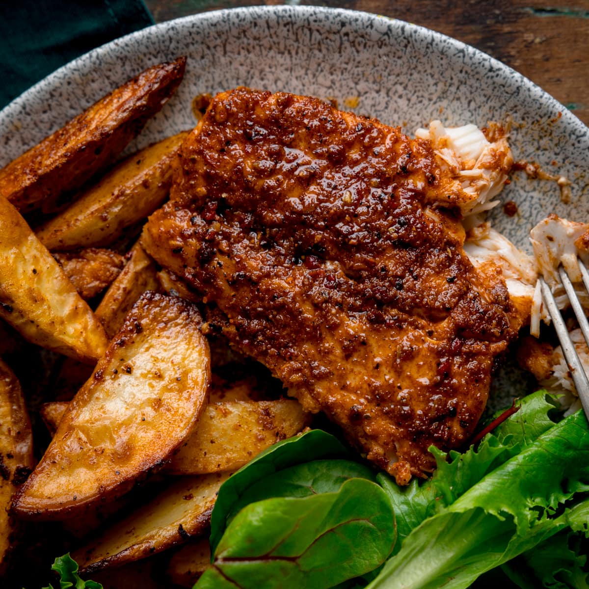 An overhead and close-up image of cajun cod, smoky potato wedges and some green salad leaves placed on a grey plate. This is on a brown background with a dark green napkin on the top left-hand side.