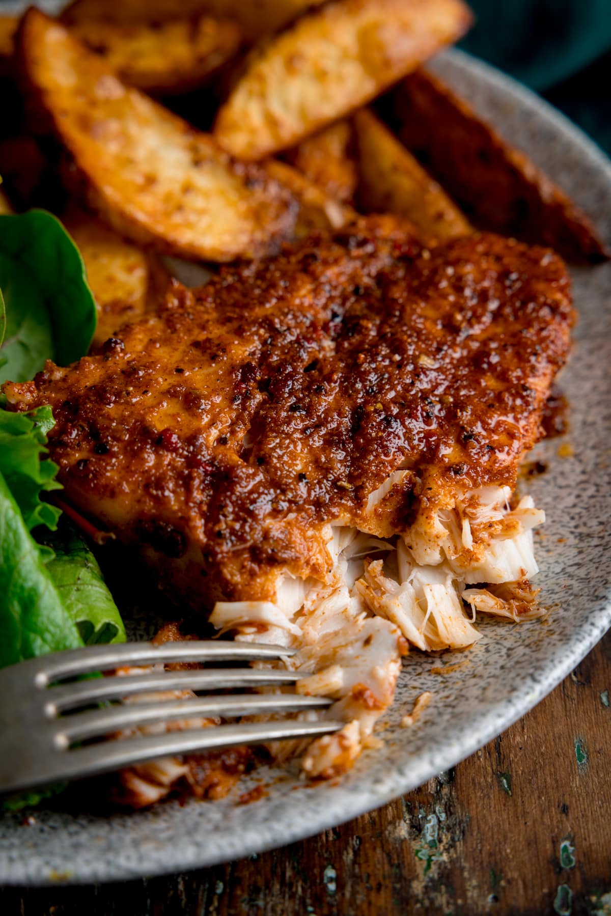 A close-up image of cajun cod, smoky potato wedges and some green salad leaves placed on a grey plate. The cod is slightly flaked with the tip of a silver fork resting on it. This is on a brown background.