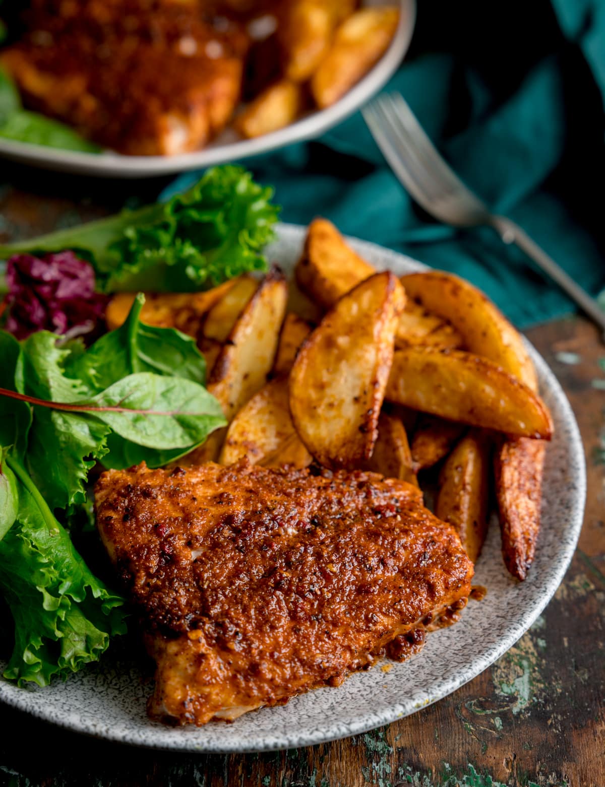 An image of cajun cod, smoky potato wedges and some green salad leaves arranged on a grey plate. This is on a brown surface. In the top left of the background is another plate identical to the main dish, on the top right is a dark green napkin with a silver fork resting on it.