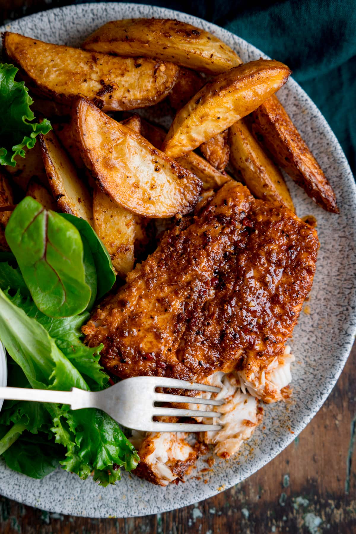An overhead image of cajun cod, smoky potato wedges and some green salad leaves placed on a grey plate. The cod is slightly flaked with the tip of a silver fork resting on it. This is on a brown background.