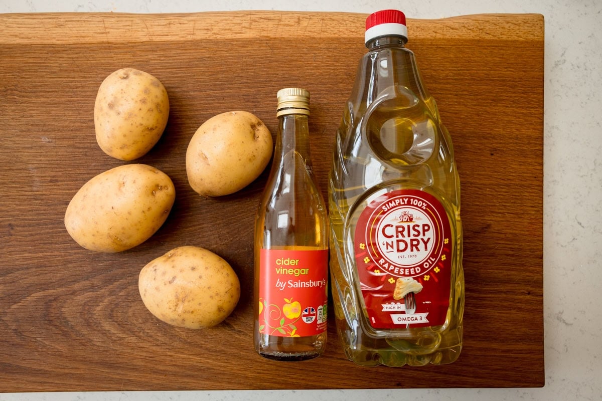 The ingredients of Chip Shop-Style Chips are laid out on a cutting board, which is on a white surface. The ingredients are: four potatoes, cider vinegar and rapeseed oil.