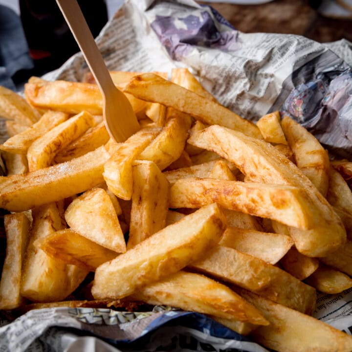 A square image of Chip Shop-Style Chips on top of some crumpled newspaper, with a small wooden fork sticking out of the chips. The chips and newspaper are placed on a wooden surface.