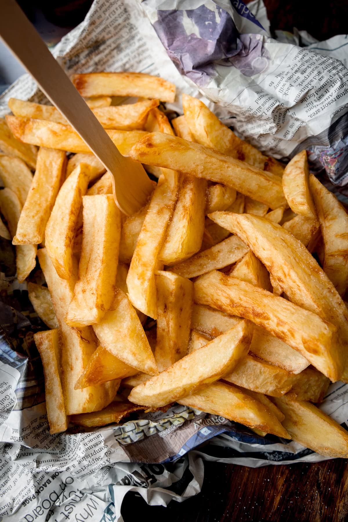 A tall, overhead image of Chip Shop-Style Chips on top of some crumpled newspaper, with a small wooden fork sticking out of the chips. The chips and newspaper are placed on a wooden surface.