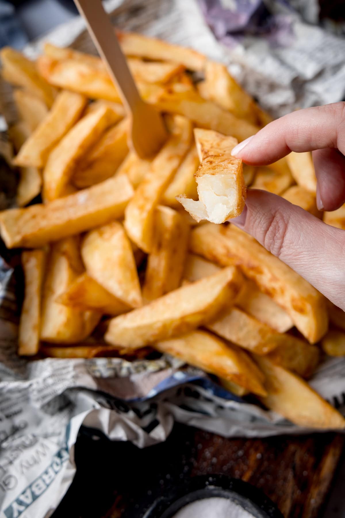 A tall, overhead image of Chip Shop-Style Chips on top of some crumpled newspaper, with a small wooden fork sticking out of the chips. There is also a thumb and forefinger holding half a chip, so the white, fluffy inside can be seen. The chips and newspaper are placed on a wooden surface. In the bottom middle of the background there is a small black dish of salt.