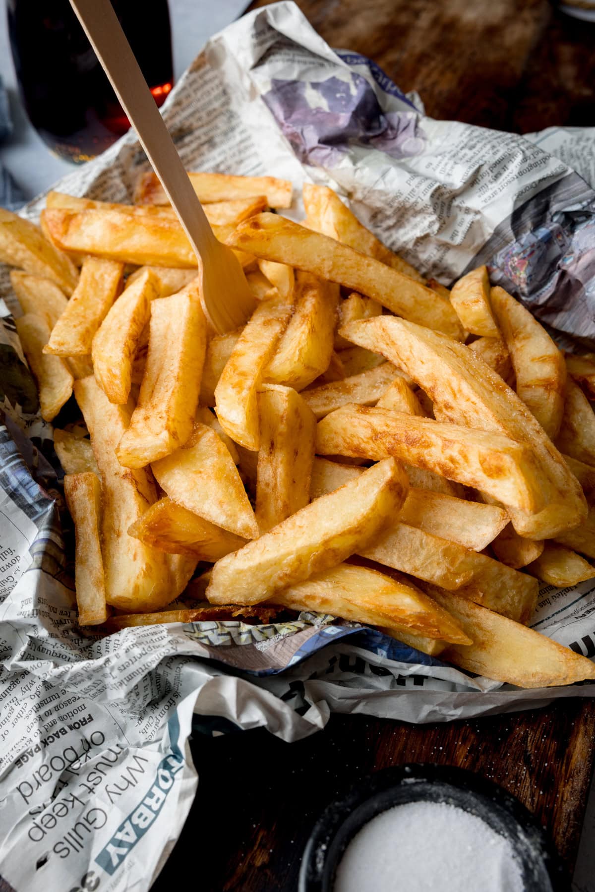 A tall, image of Chip Shop-Style Chips on top of some crumpled newspaper, with a small wooden fork sticking out of the chips. The chips and newspaper are placed on a wooden surface. In the bottom middle of the background, there is a small black dish of salt. And, in the top left of the background, you can slightly see a brown vinegar bottle.