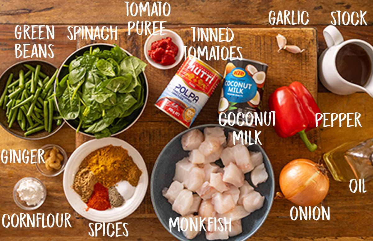 An overhead shot of the ingredients in Monkfish Coconut Curry laid out on a wooden board. The ingredients are labelled in white, they are as follows: tomato puree, tinned tomatoes, coconut milk, monkfish, spices, cornflour, ginger, green beans, spinach.