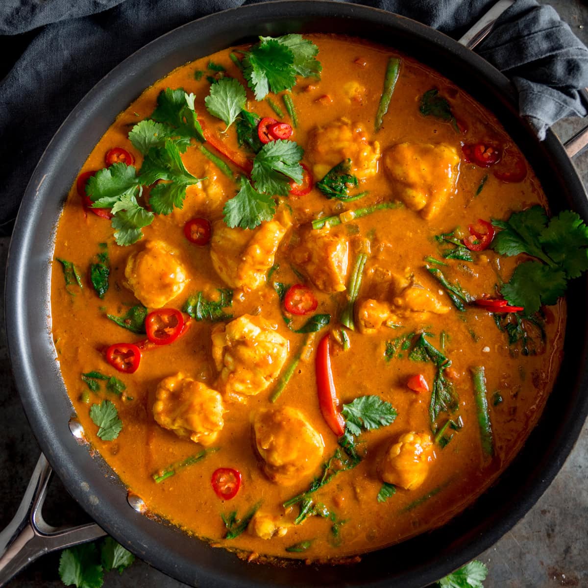 A square overhead shot of Monkfish Coconut Curry in a grey pan, topped with coriander. On a grey surface with a grey napkin in the background.