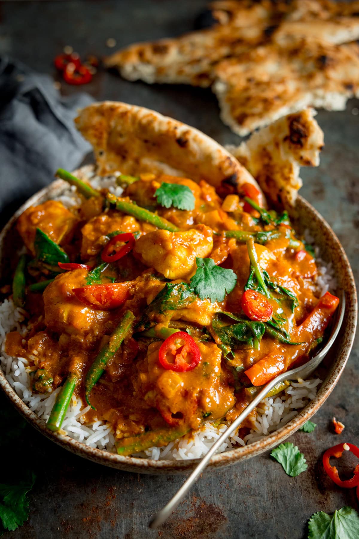 Monkfish Coconut Curry on top of rice in a grey/brown bowl, topped with coriander and red chillies. There is a fork sticking out the bottom of the bowl and a couple of pieces of naan in the curry. This in on a grey background with a grey napkin. There is a piece of naan in the background as well as coriander scattered around.