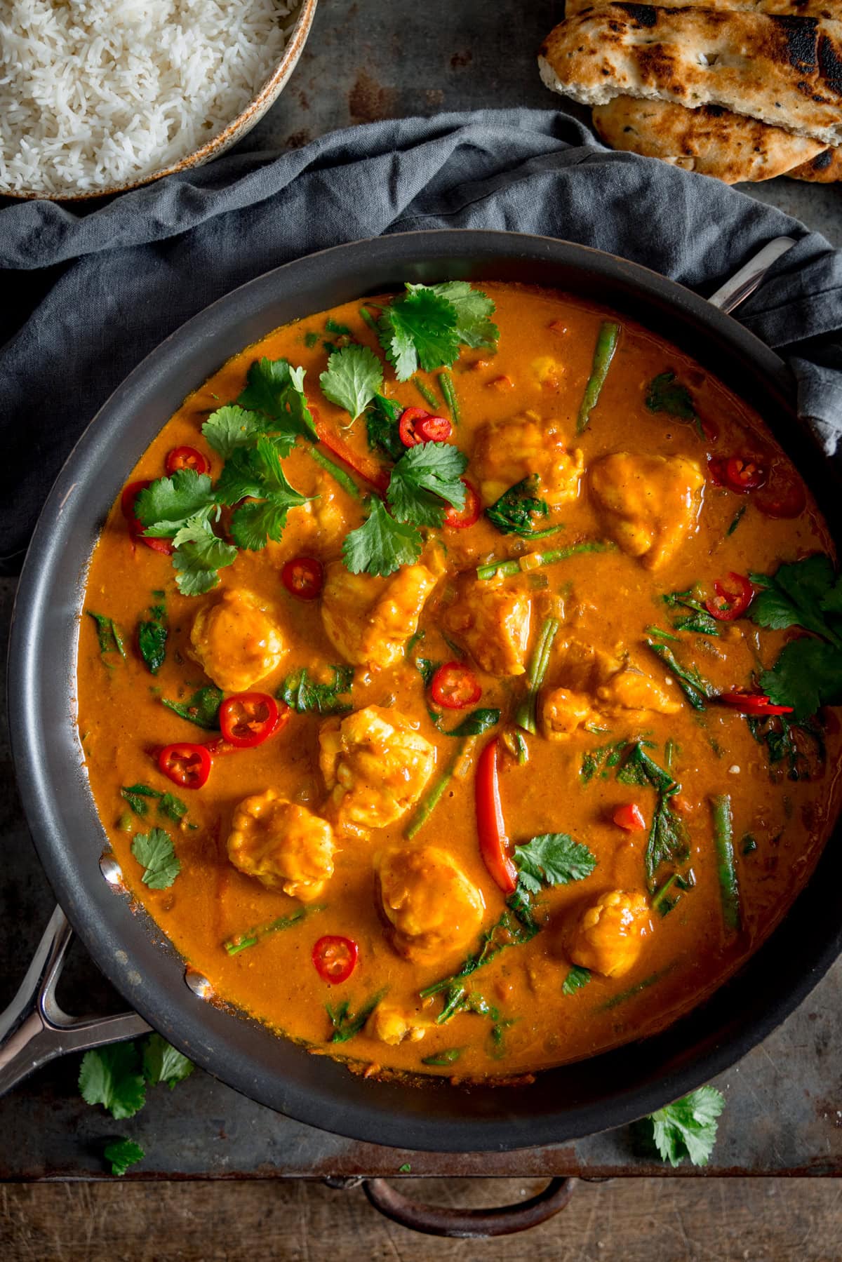 A tall overhead shot of Monkfish Coconut Curry in a grey pan, topped with coriander. In the background on the top left there is a grey bowl filled with rice, and on the top righ there is 2 naan On a grey surface with a grey napkin in the background.