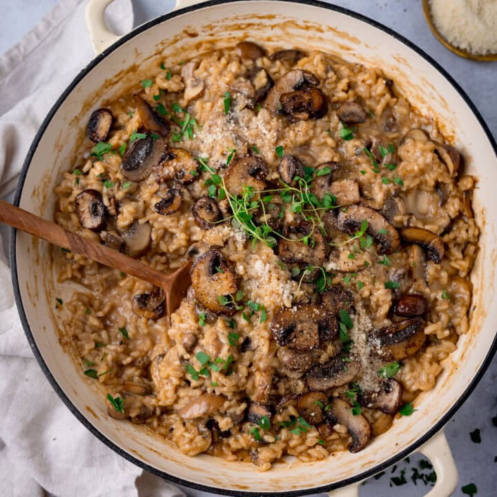 A square image of Mushroom Risotto in a white/cream-coloured crock pot. There are some sprigs of thyme on top of the risotto. There is a wooden spoon sticking out of the dish. On the left of the dish there is a cream-coloured napkin tucked beside the crock pot. In the right of the background, there is some dried parsley scattered around. On the top right, there is a gold dish of grated parmesan. This is all on a light grey background.
