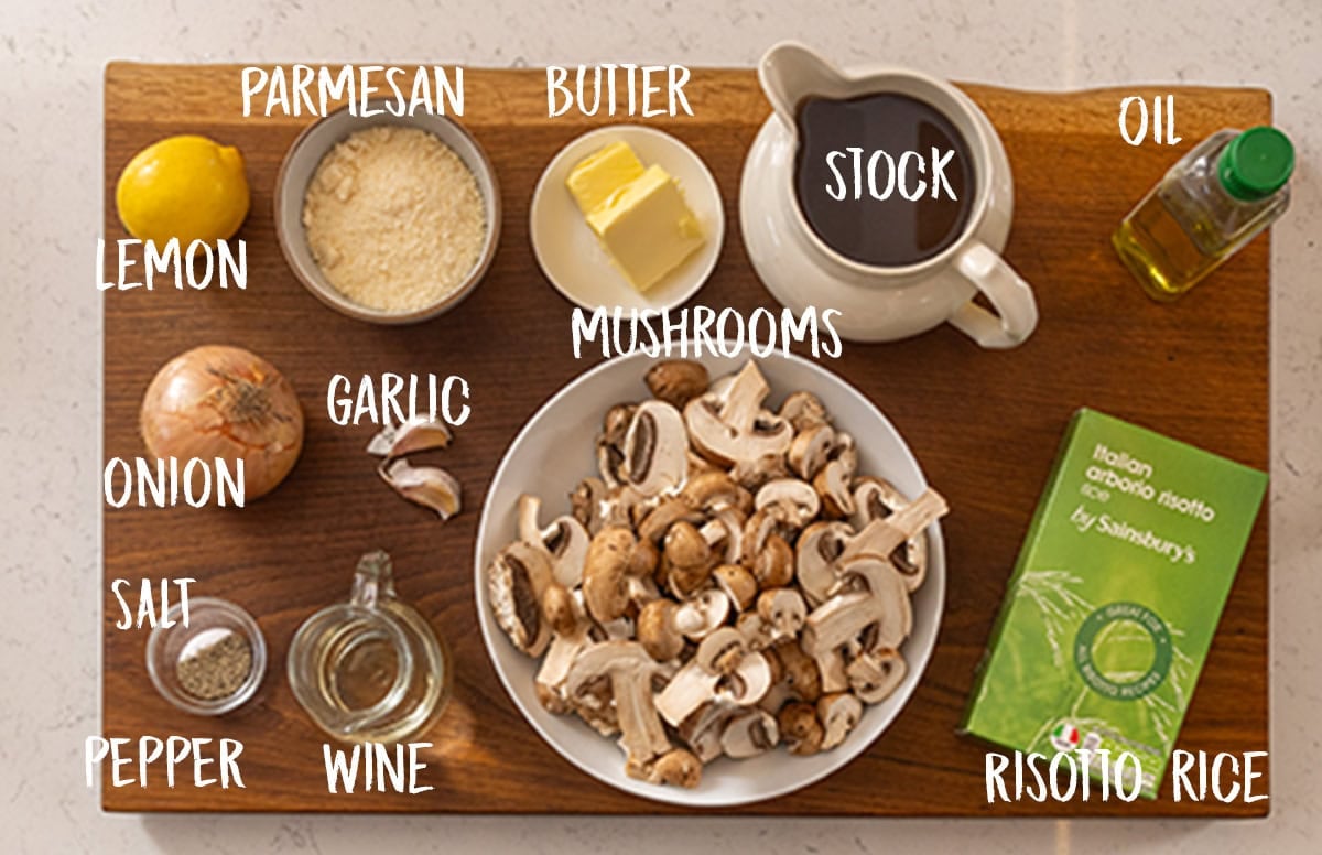 The ingredients for Mushroom Risotto are laid out on a wooden board, which is placed on a white marble surface. They are as follows: lemon, onion, salt, pepper, parmesan, garlic, wine, butter, mushrooms, stock, oil, risotto rice.