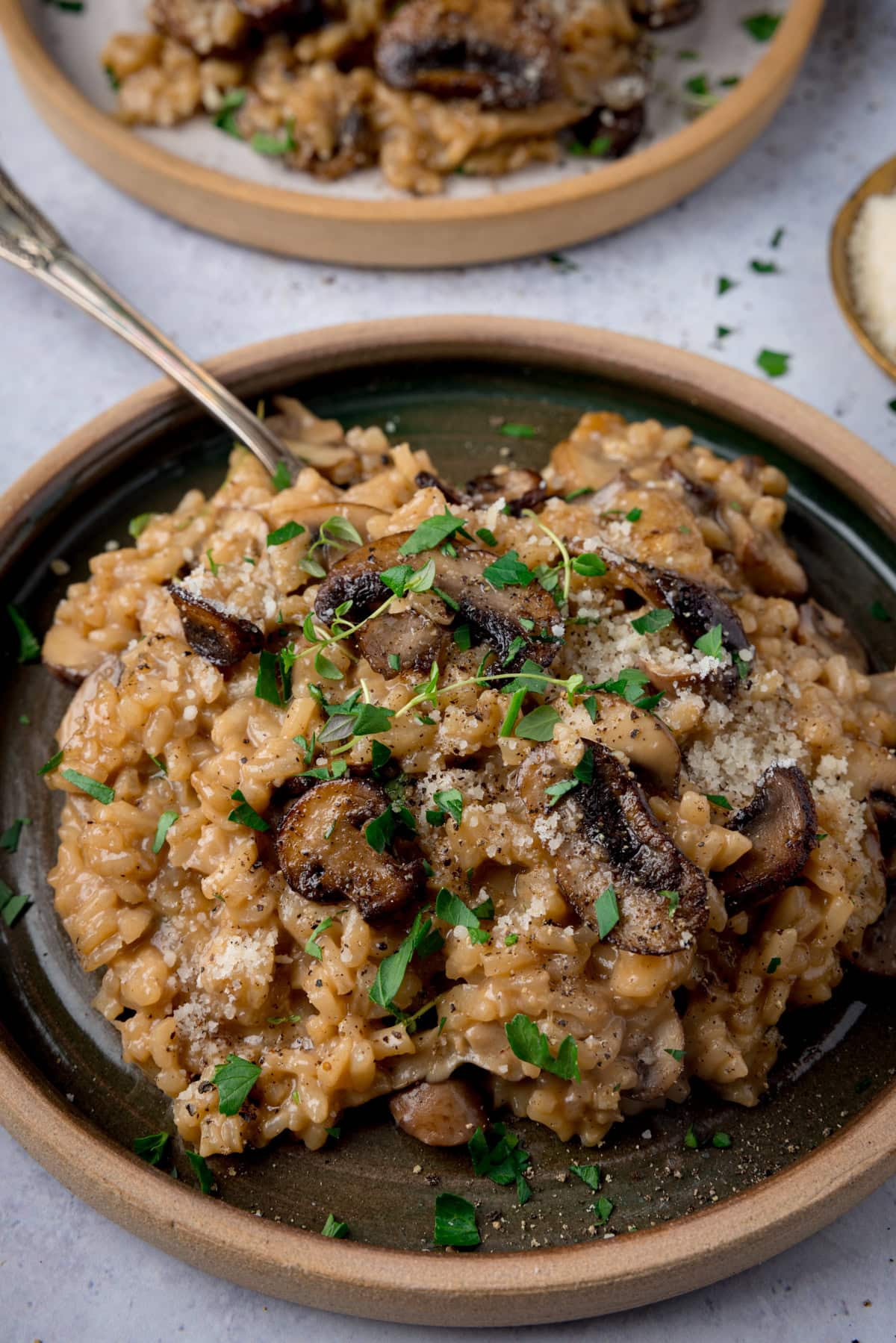 A tall image of Mushroom Risotto. In the centre of the image there is a dark green dish with a brown rim, which contains a portion of Mushroom Risotto, topped with sprigs of thyme, parmesan and ground black pepper. There is a silver fork sticking out of the risotto. In the top of the background there is another plate of Mushroom Risotto, except this plate is white with a brown rim. On the right of the background, there is a gold dish of parmesan, with some parsley sprinkled around it. This is on a light grey surface.