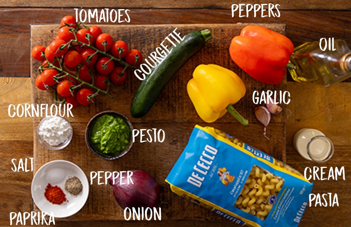 The ingredients of Roasted Vegetable Pesto Pasta laid out on a wooden cutting board, which is on a wooden surface. The ingredients are labelled in white text. They are as follows: tomatoes, courgette, peppers, garlic, oil, cream, pasta, onion, pesto, salt, pepper, paprika, and cornflour.