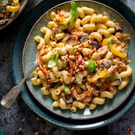 A square overhead image of roasted vegetable pesto pasta topped with some small basil leaves and roasted pine nuts, on a light blue plate, which is placed on a larger dark blue plate. There is a silver fork sticking out of the pasta. In the top left corner of the image, you can see the corner of a light blue dish with more roasted vegetable pesto pasta. There are some roasted pine nuts scattered around the background, which is grey.
