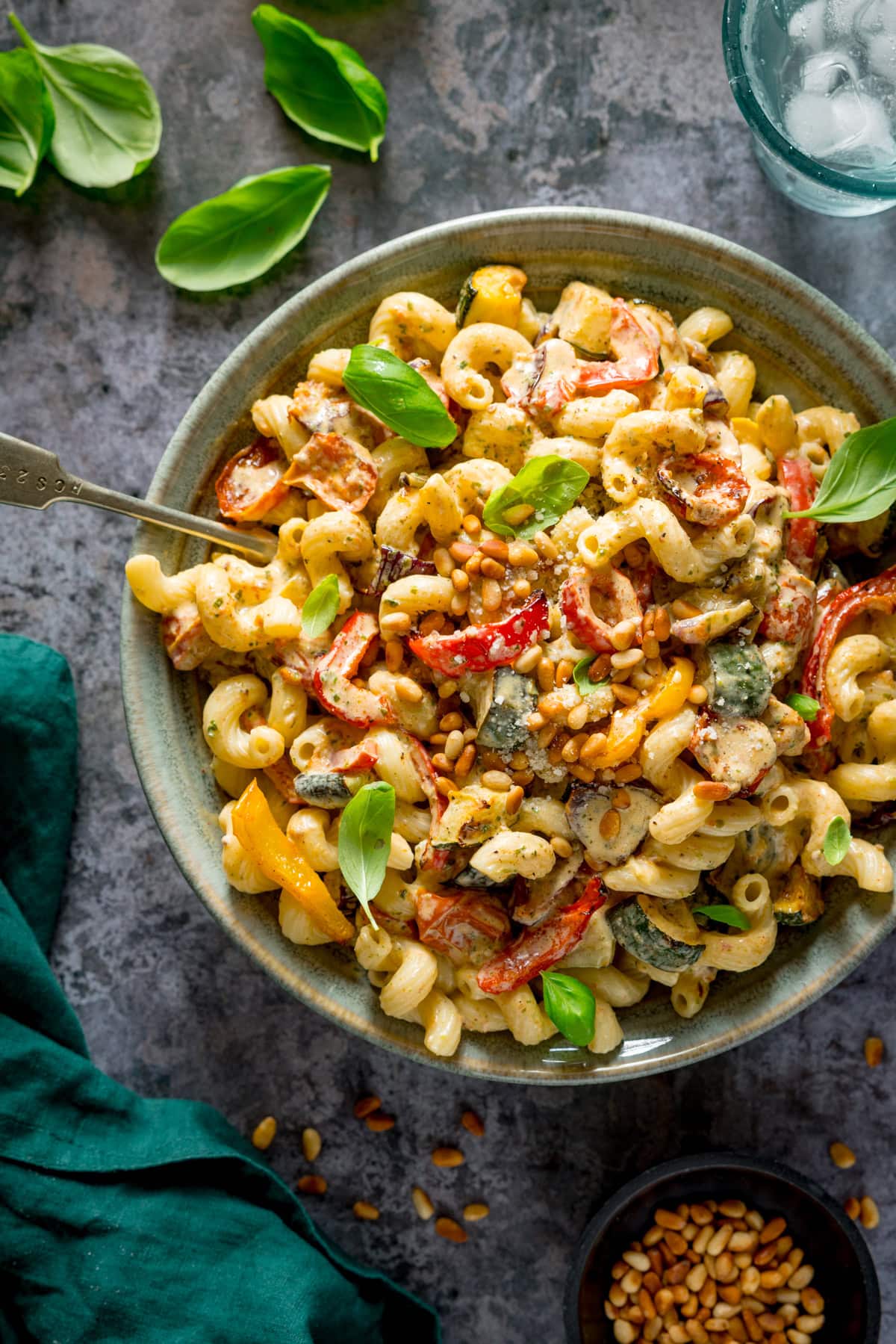 A tall, overhead image of roasted vegetable pesto pasta, topped with small basil leaves, and some toasted pine nuts, in a light blue/grey bowl, with a silver fork sticking out of the pasta. On the top right of the image, there is a glass of water with ice, on the top left there are a few medium-sized basil leaves scattered around. On the bottom right there is a small black dish of toasted pine nuts, with more toasted pine nuts scattered around. On the bottom left, there is a teal/green napkin. This is all placed on a grey background.