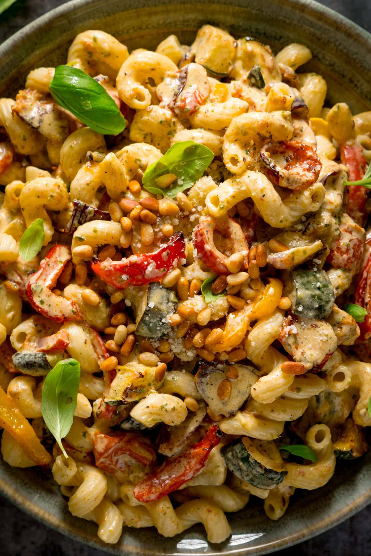A very close-up tall image of roasted vegetable pesto pasta topped with toasted pine nuts, and a few small basil leaves. This is in a light grey bowl. The pasta an the bowl fill the whole image, so nothing else can be seen.