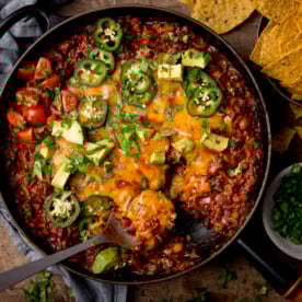 Tex-Mex minced beef in a black cast iron pan with cheese, jalapenos, coriander and avocados on top. There is a silver spoon sticking out of the dish. There is a grey dish of coriander on the bottom right of the image, a grey napkin on the top left and some tortilla chips on the top right. This is on a wooden background.