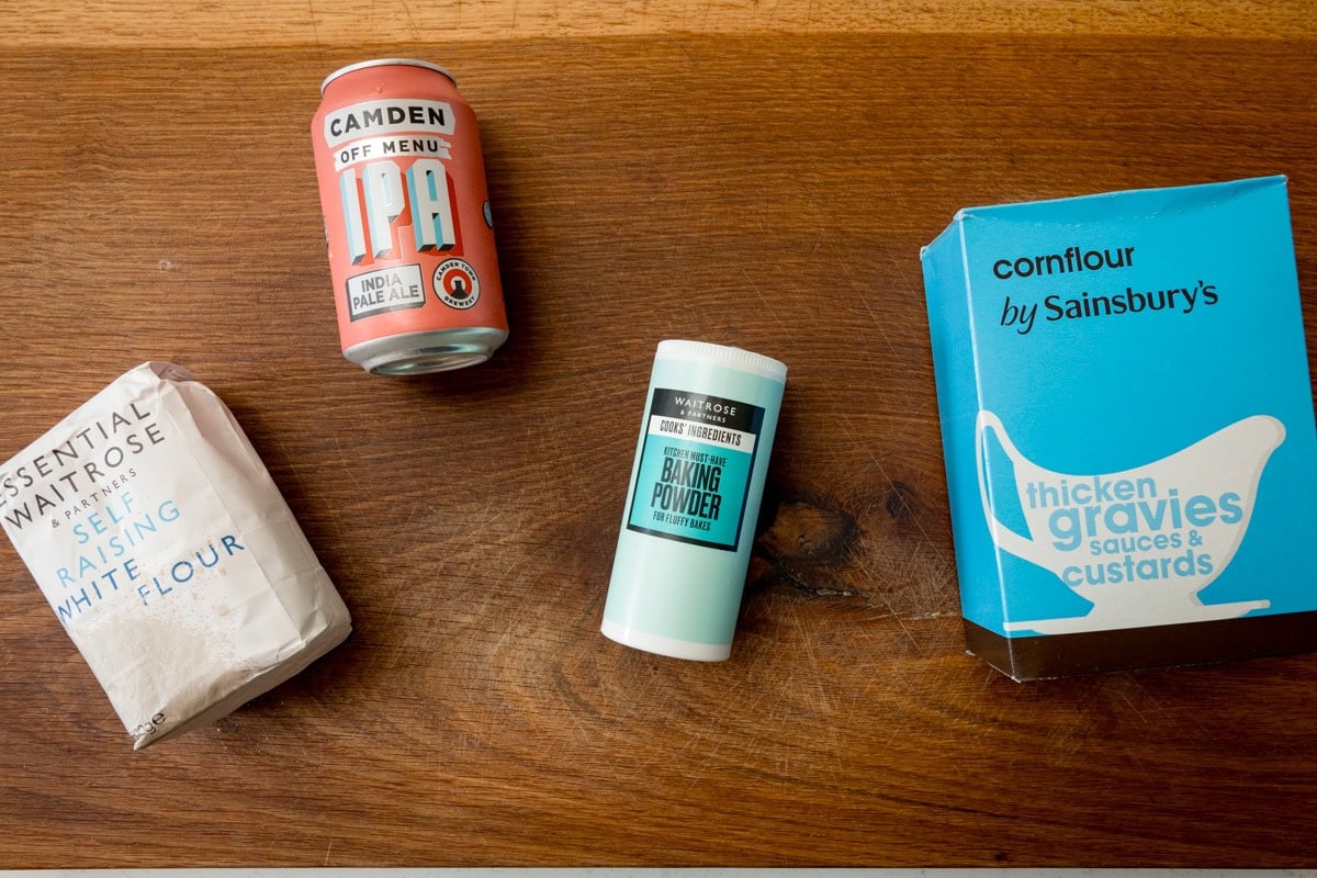 An overhead shot of the ingredients for the beer batter is laid out on a wooden board. They are as follows: self-raising white flour, IPA (beer), baking powder, and cornflour.