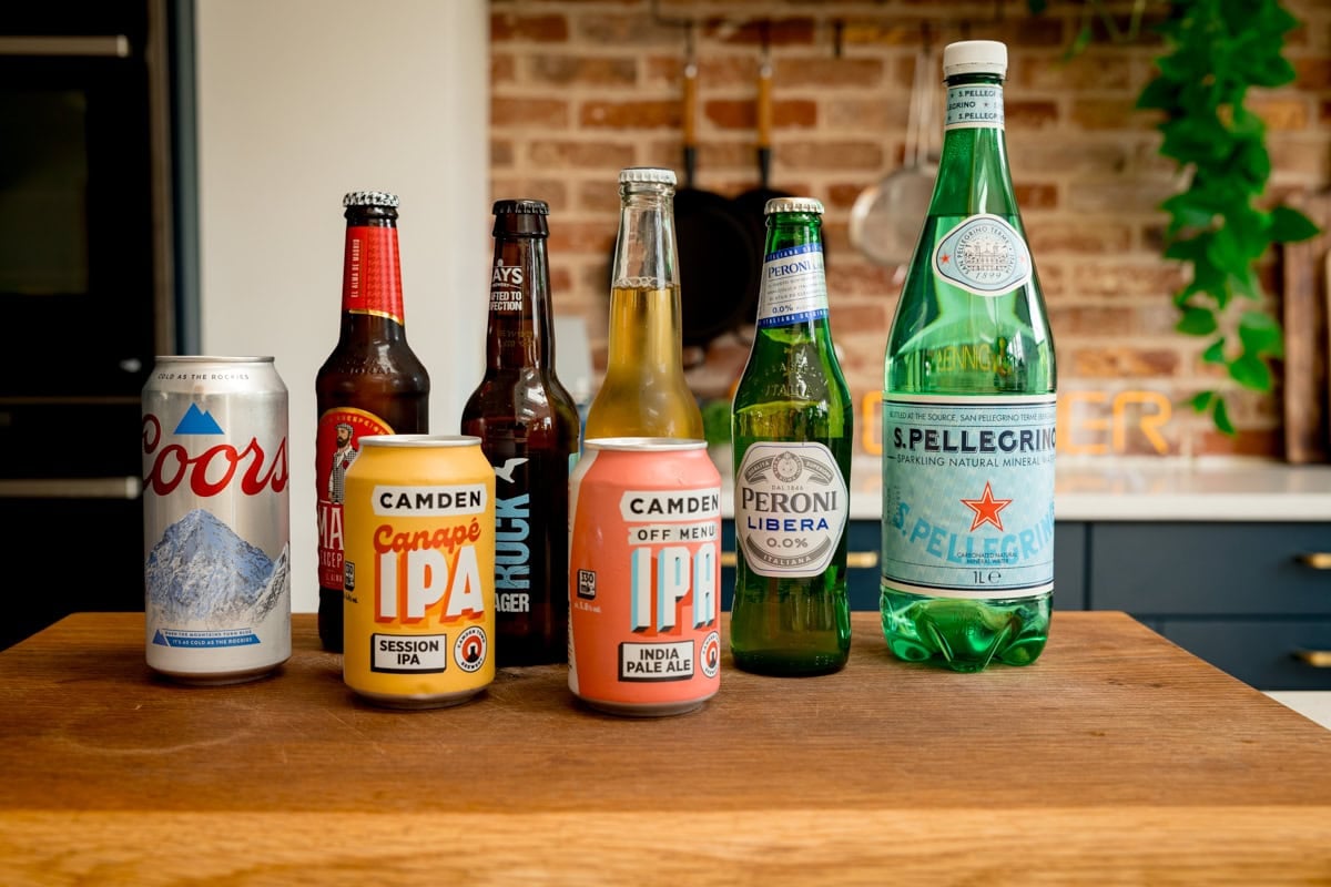 A wide image of 8 bottles and cans, standing grouped together on top of a wooden cutting board. They are the liquid options you can use for beer batter. They are as follows: pale ales, beer, larger, 0% alcohol beer, and san Pellegrino sparkling water.