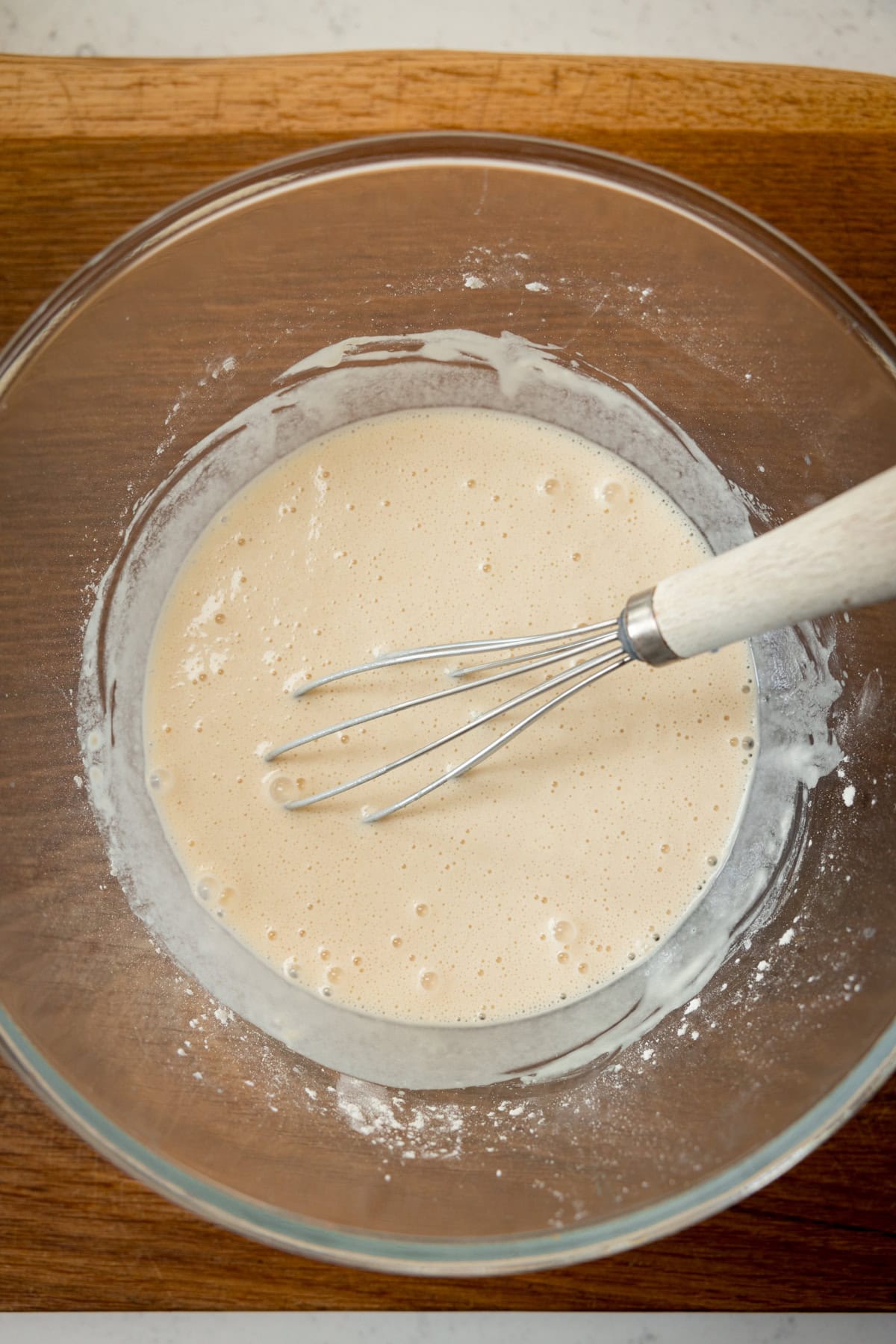 A tall, overhead shot of the finished beer batter. The batter is in a clear, glass bowl with a whisk wooden handle in the batter. All this is laid on a wooden board.