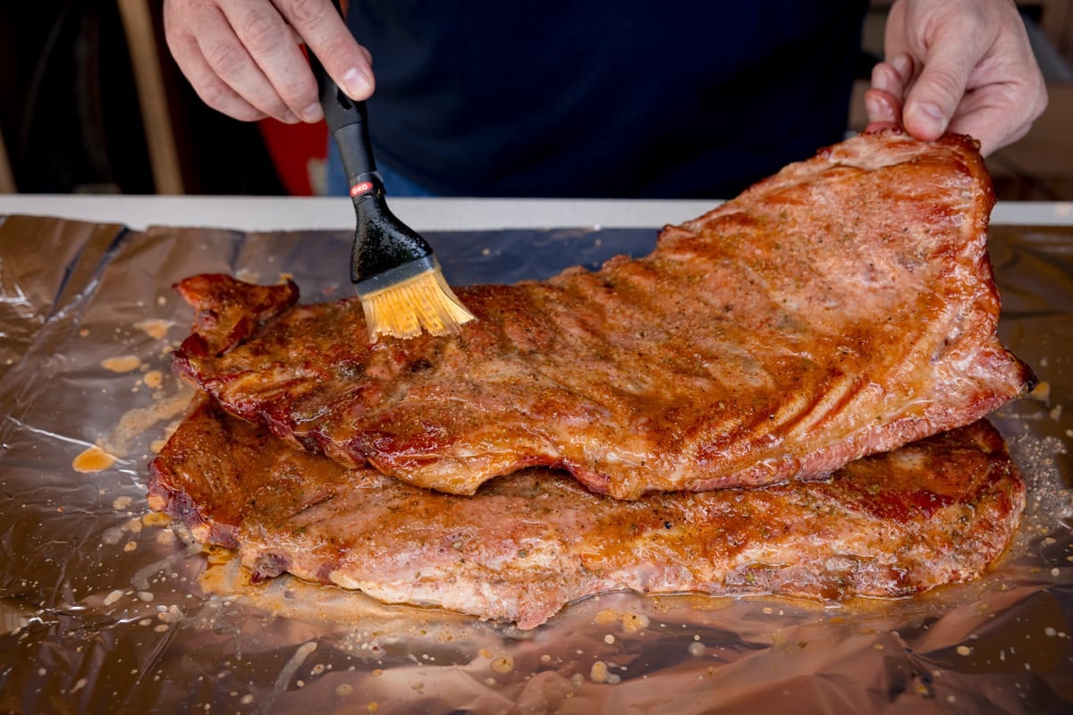 A prep shot of BBQ Dry Rub Ribs. The ribs are laid out on a sheet of silver tinfoil, which is on a white surface. There are two hands visible, one is holding one end of the ribs, and the other is holding a brush and applying the mop.