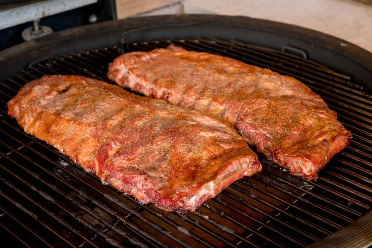 A prep shot of BBQ Dry Rub Ribs. There are two racks of smoked and seasoned ribs laid out on a black barbeque, which is heated to 120 degrees Celsius. The ribs are slightly more golden.