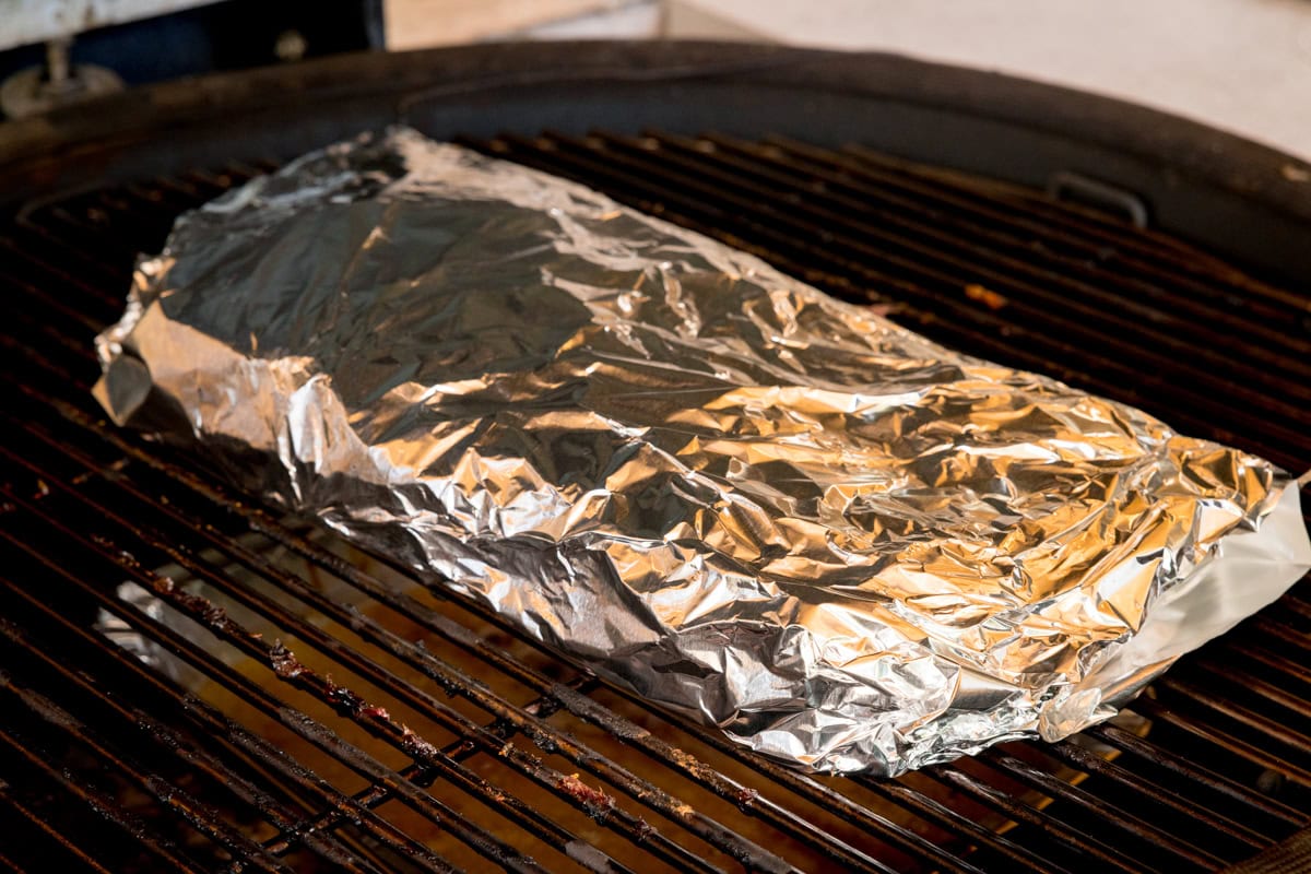 A prep shot of BBQ Dry Rub Ribs. The ribs are wrapped in silver tin foil and placed on a black barbeque, which is heated to 120 degrees Celsius.