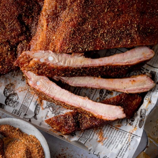 A square, close-up image of dry rub BBQ ribs, there are two racks of the ribs laid out on a silver tray that is lined with newspaper. The central rack of ribs is cut into and the three of the ribs are separated so you can see the ribs. In the bottom left of the background, there is the dry rub seasoning in a light grey bowl with a silver teaspoon sticking out of it. On the bottom right of the image, there is a large silver knife with a wooden handle set at an angle. This is all set on a light grey background.