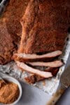 A tall, close-up image of dry rub BBQ ribs, there are two racks of the ribs laid out on a silver tray that is lined with newspaper. The central rack of ribs is cut into and the three of the ribs are separated so you can see the ribs. In the bottom left of the background, there is the dry rub seasoning in a light grey bowl with a silver teaspoon sticking out of it. On the bottom right of the image, there is a large silver knife with a wooden handle set at an angle. This is all set on a light grey background.