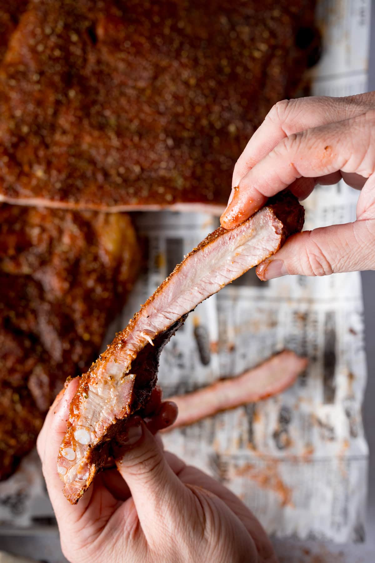 A tall, close-up image of dry rub BBQ ribs, there are two racks of the ribs laid out on a silver tray that is lined with newspaper. The central rack of ribs is cut into and the two of the ribs are separated so you can see the ribs. In the foreground there are two hands picking up one rib, showing the inside of the rib to the camera. This is all set on a light grey background.